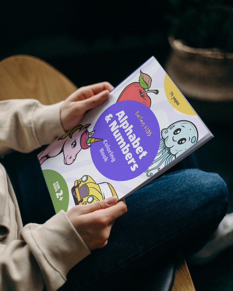 A person holding a children’s ‘Alphabet & Numbers Coloring Book’ by Curious Kids, featuring colorful illustrations of an apple, jellyfish, unicorn, and yellow car.